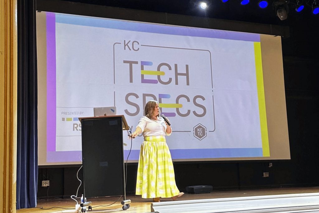 A presenter speaks into a handheld microphone at a lectern, gesturing toward a large projected slide behind her on a stage in an auditorium.

Transcribed text on slide: "KC TECH SPECS" 
Also visible: "PRESENTED BY:" and a small hexagon logo reading "KC TC"