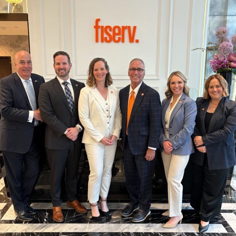 Six professionally dressed adults stand smiling and posing in a corporate lobby, marble floor beneath them, floral arrangement to the side, with an orange sign reading "fiserv." on the wall.
