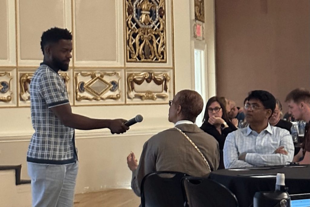 A standing man extends a microphone to a seated woman speaking, while other attendees listen at round tables in an ornate conference room. Text in image: "EXIT".