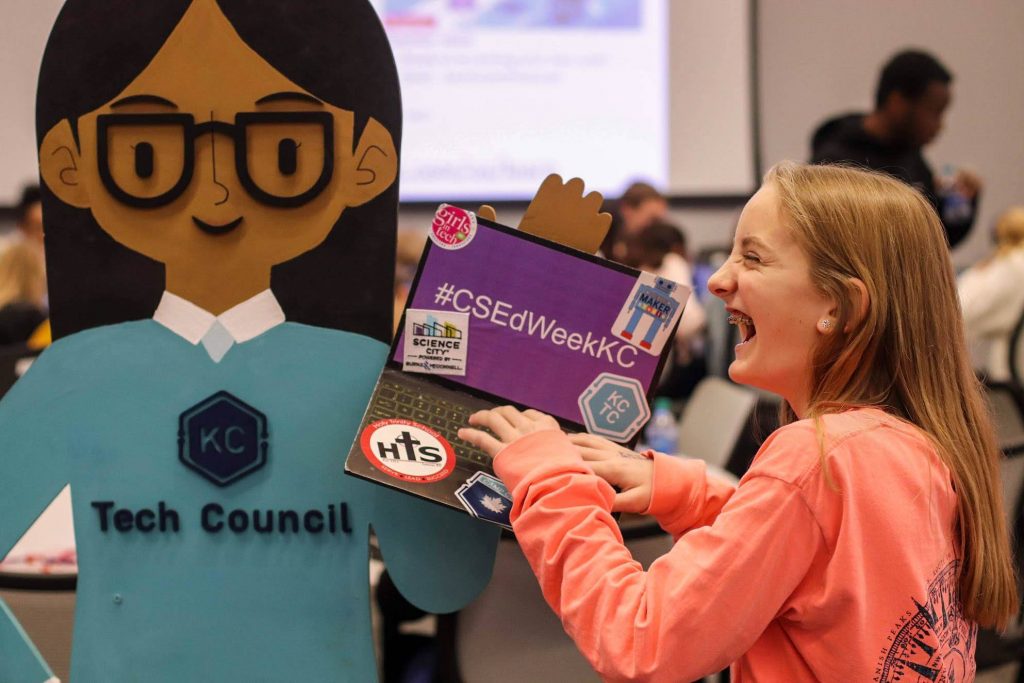 Cardboard figure holding a stickered laptop while a smiling girl laughs and pretends to type in a busy conference room.

Text visible: #CSEdWeekKC; Tech Council; KC; Science City; MAKER; HTS