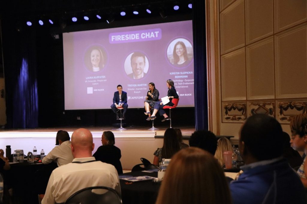 Projection screen displays a panel; three seated panelists converse on stools at front while an audience watches in a dim conference hall.

Legible on-screen text: "FIRESIDE CHAT" ; "TREVOR MARTIN" ; "H&R BLOCK". (Other smaller text and names are present on the slide but are not fully legible in the photo.)