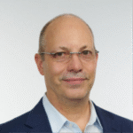 Middle-aged man with glasses and receding hairline, smiling slightly while facing the camera; wearing a navy blazer and light-blue collared shirt against a plain light-gray background.