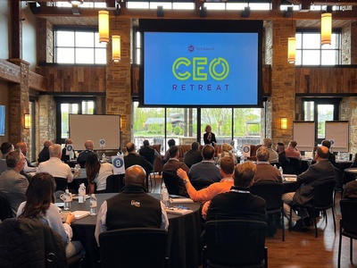 Large projection screen displays "CEO RETREAT" while a presenter addresses seated attendees in a rustic conference room with round tables and visible table signs reading "8".

Transcribed text: CEO RETREAT; 8