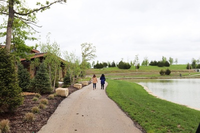 Two people walk away along a paved path beside a pond, flanked by grass, young trees and a small wooden building under an overcast sky.
