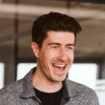 Smiling man with short dark hair and stubble, wearing a gray patterned shirt over a black tee, laughing in a bright office space with glass partitions and soft overhead lighting.
