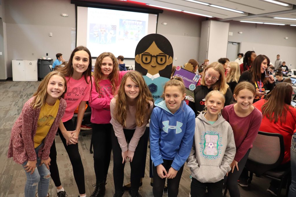 Group of smiling school-age girls pose around a cardboard cartoon character, standing in a busy tech classroom with students at laptops and a projection screen in the background.

Text in image: #CSEdWeekKC; KC (on cardboard badge).