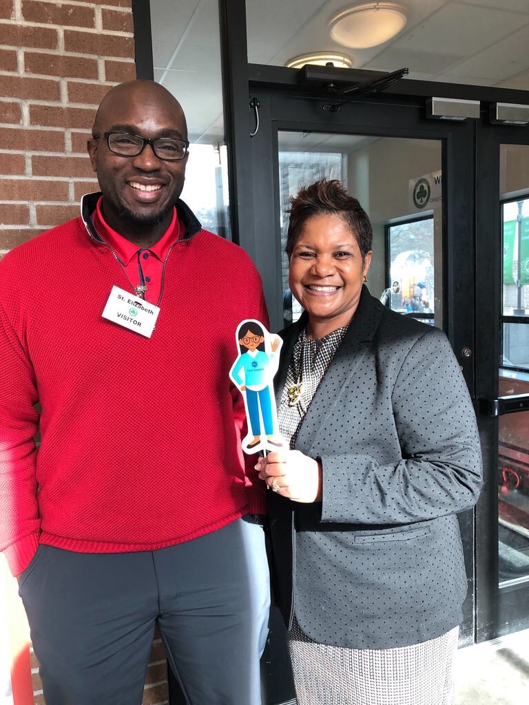 A man and a woman smile at the camera; the woman holds a small cartoon cutout. They stand together by a brick wall and glass entry doors in a lobby.

Transcribed text: "St. Elizabeth" ; "VISITOR"
