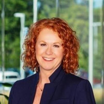 Woman with curly red hair smiling at the camera, wearing a dark blazer, posed indoors before large glass windows showing blurred green trees and parked cars outside.